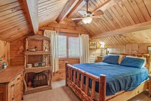 A wooden cabin bedroom with a bed, a shelf with items, a window, and a ceiling fan.