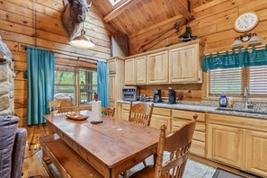 A wooden cabin with a dining table, chairs, a kitchen with appliances, and a clock on the wall.
