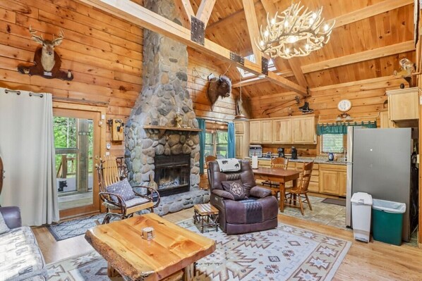 A log cabin interior with a stone fireplace, wooden furniture, a dining area, and a kitchen.