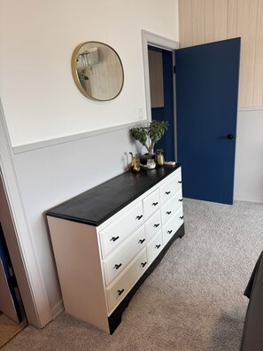A white dresser with black top, a round mirror, a potted plant, and a blue door in a room with beige carpet.