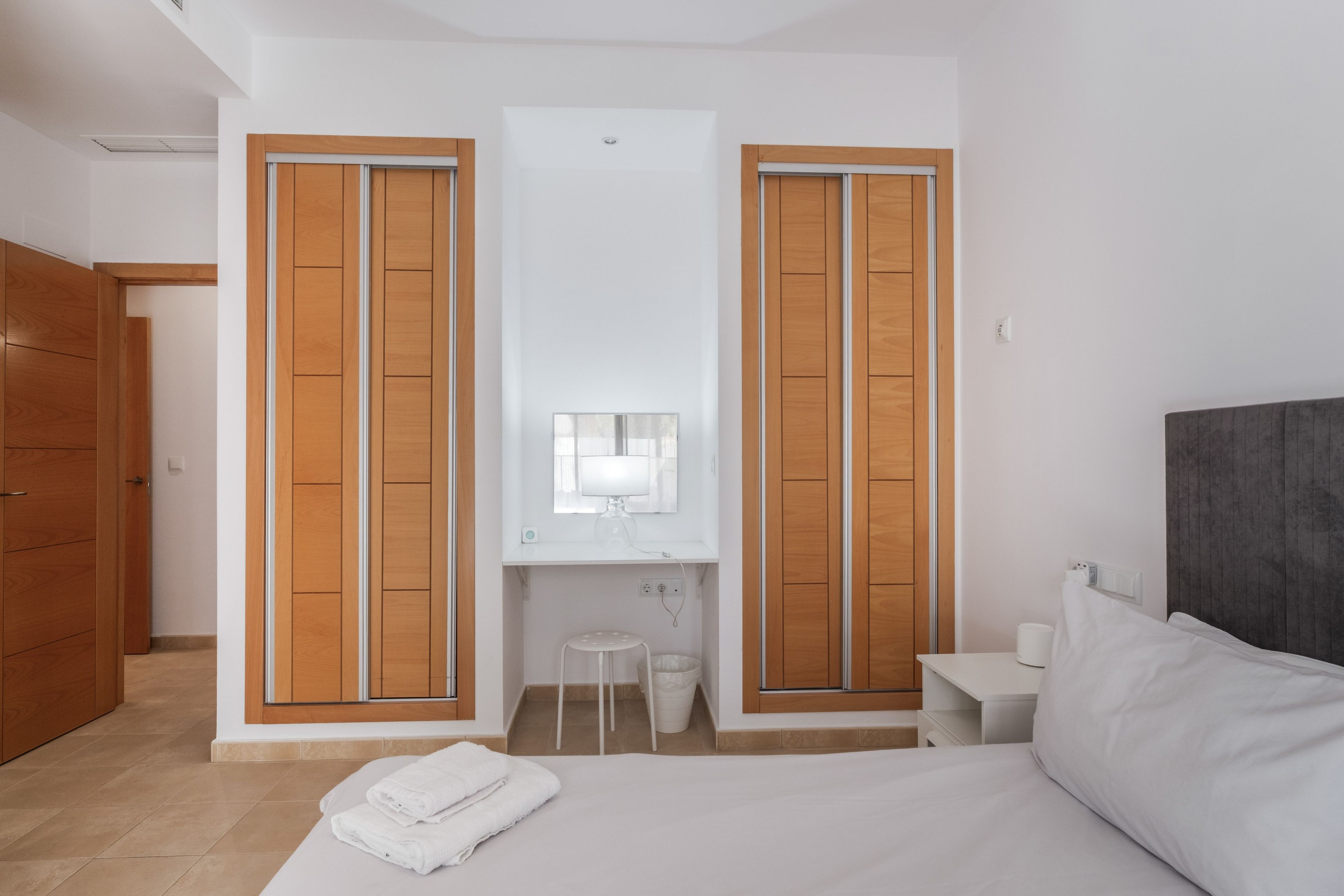 Een hotelkamer met een bed, een klein krukje, een nachtkastje en een spiegel boven een wastafel.