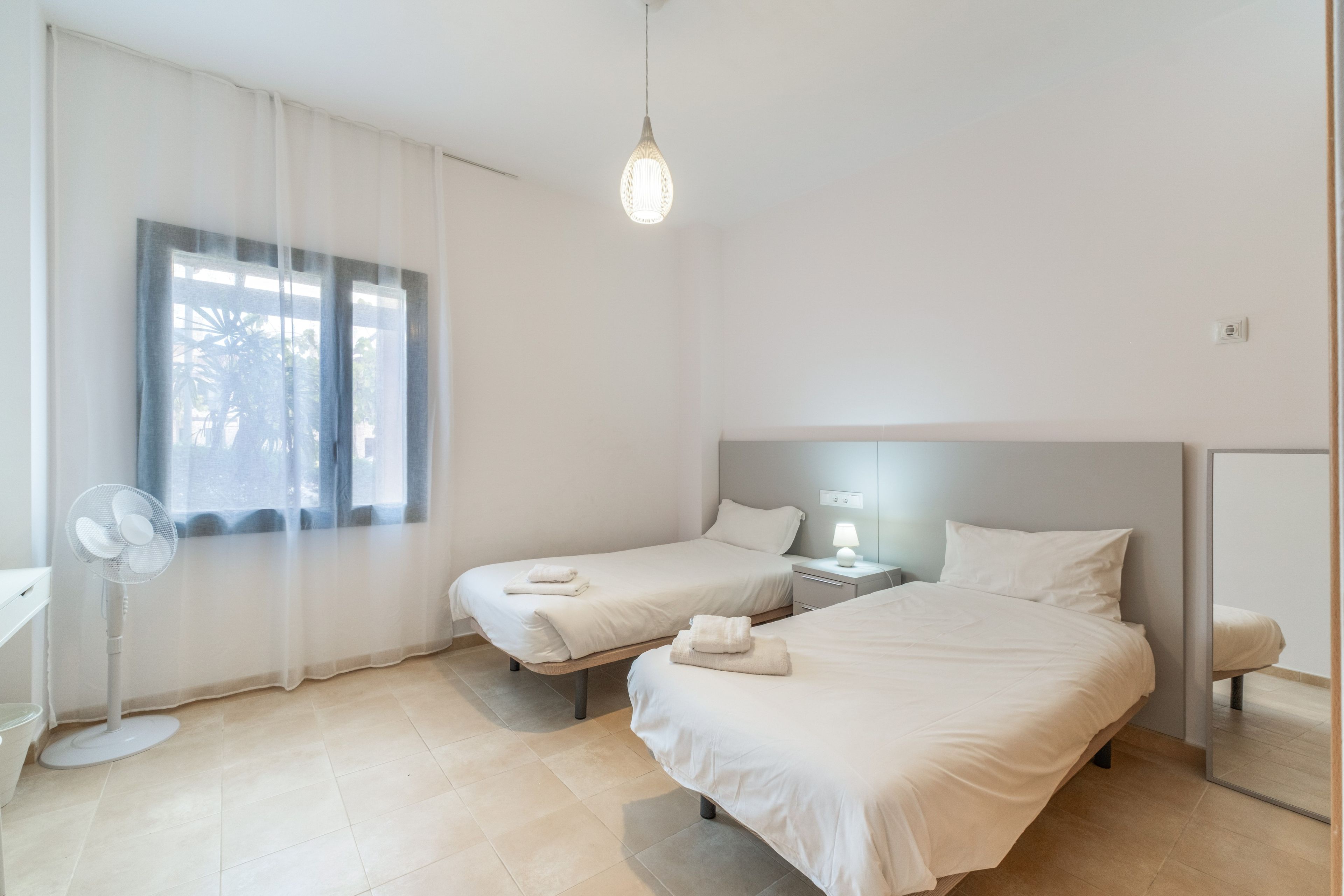 Habitación de hotel con dos camas, una mesita de noche con lámpara, un ventilador y una ventana con cortinas traslúcidas.