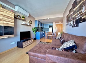 A modern living room with a brown sofa, a flat-screen TV, and a kitchen in the background. - MAENA Esmée - 🌴Appart` de Charme - Vue Marina, Clim & Piscine (Roses)