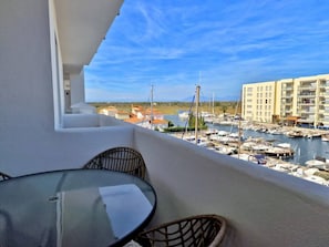 A balcony with a glass table and wicker chairs, overlooking a marina with boats and a building in the background. - MAENA Esmée - 🌴Appart` de Charme - Vue Marina, Clim & Piscine (Roses)
