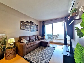 A modern living room with a sectional sofa, a patterned rug, a flat-screen TV, and a balcony with outdoor seating. - MAENA Esmée - 🌴Appart` de Charme - Vue Marina, Clim & Piscine (Roses)