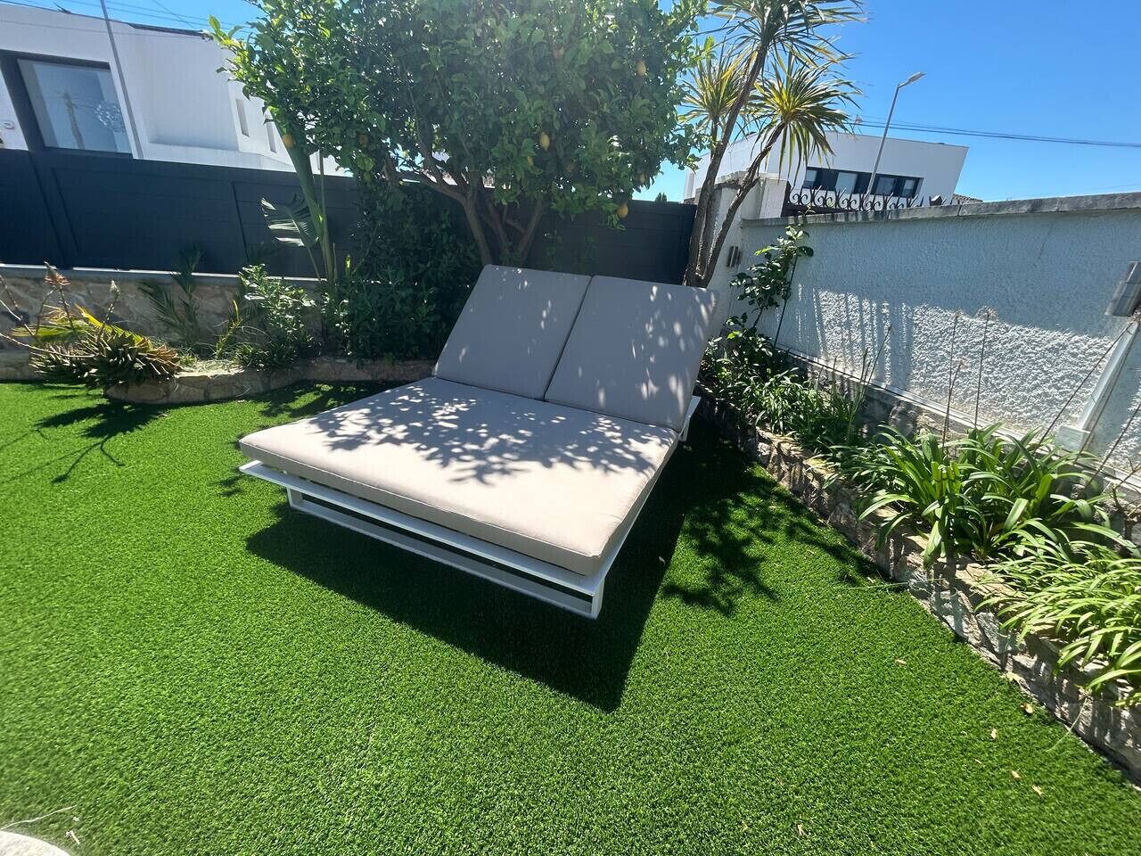 Ein Bett mit weißer Matratze und grauem Kopfteil, umgeben von grünem Kunstgras, mit einem Baum und Pflanzen in der Nähe.