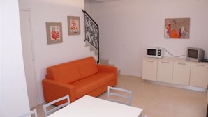 Ein kleiner, heller Raum mit einem weißen Tisch, einem orangefarbenen Sofa und einer Küchenzeile mit Mikrowelle und Fernseher.