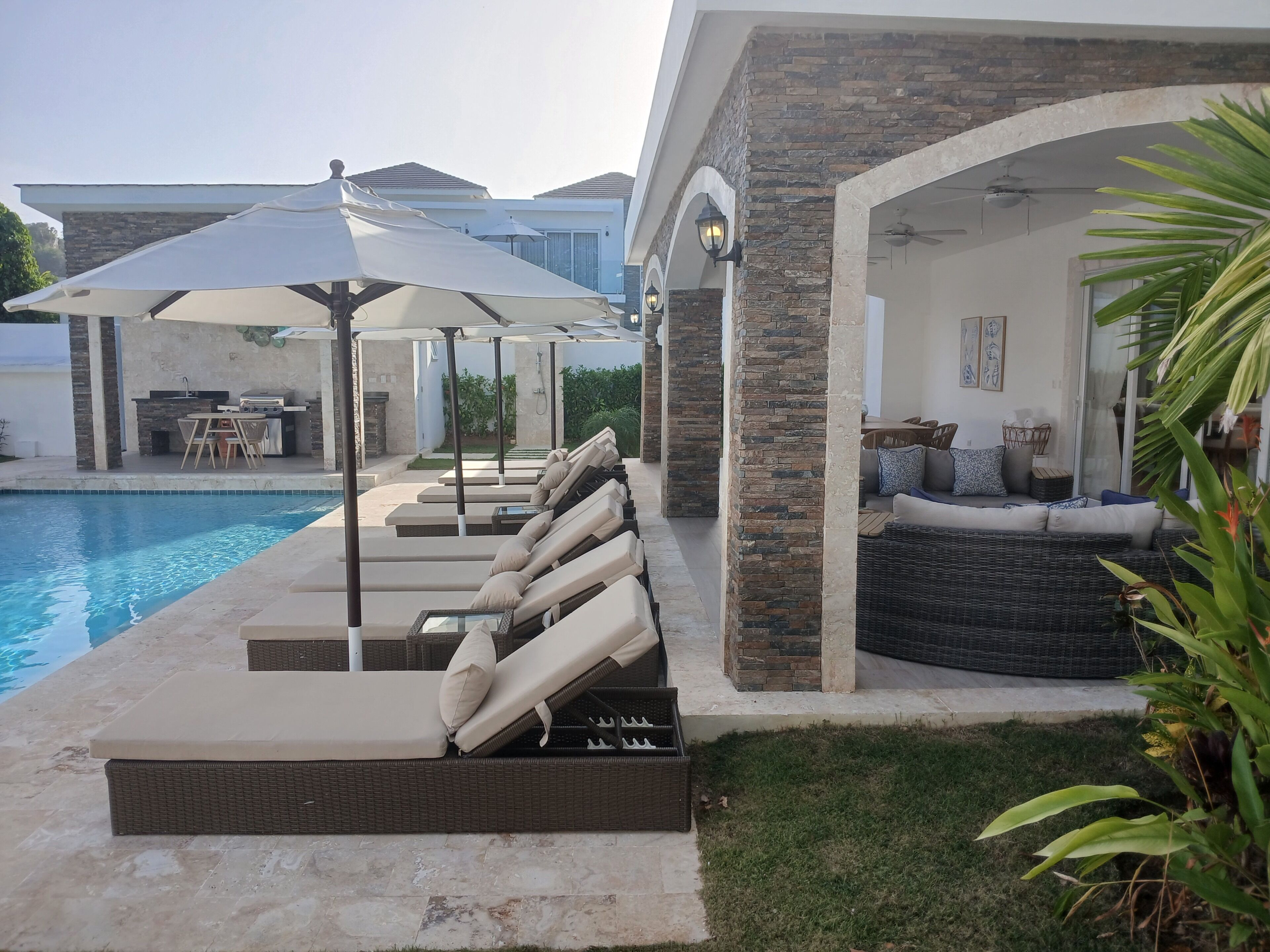 A poolside area with lounge chairs and umbrellas, a barbecue setup, and a covered seating area with cushions.