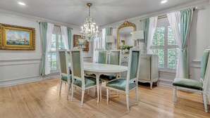 Une salle à manger avec une table blanche, des chaises recouvertes d’un tissu vert, un lustre et un grand miroir.