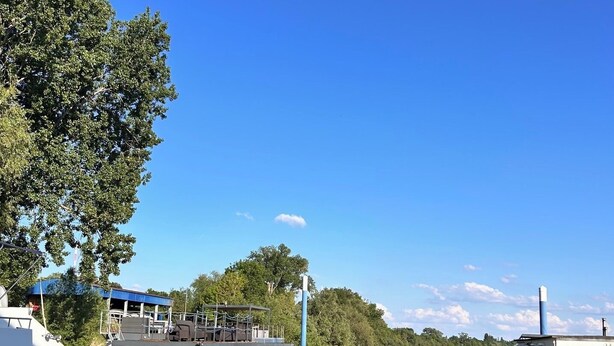 Une rivière paisible où sont amarrés des bateaux, un espace abrité avec des sièges, et un bâtiment doté d’une cheminée.