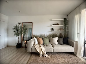 Un salon avec un canapé beige, un miroir, des étagères ornées d’objets décoratifs et une plante en pot.