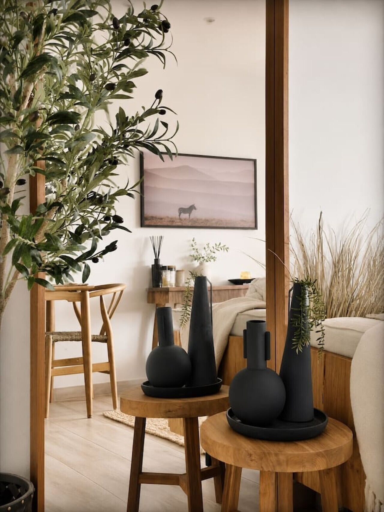 A room with a wooden floor, a wooden chair, a round wooden table with black vases, a wall-mounted picture of a horse, and a large plant.