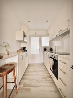 Une cuisine étroite avec des meubles blancs, un tabouret en bois et une vue sur l’extérieur, visible à travers une porte.