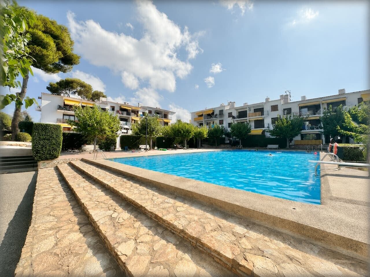 Une piscine accessible par des marches, entourée de bâtiments dotés de balcons, sous un ciel dégagé.