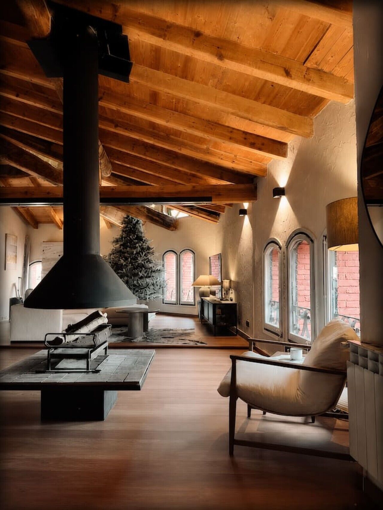 Acogedor loft con chimenea grande, vigas de madera y ventanas arqueadas.