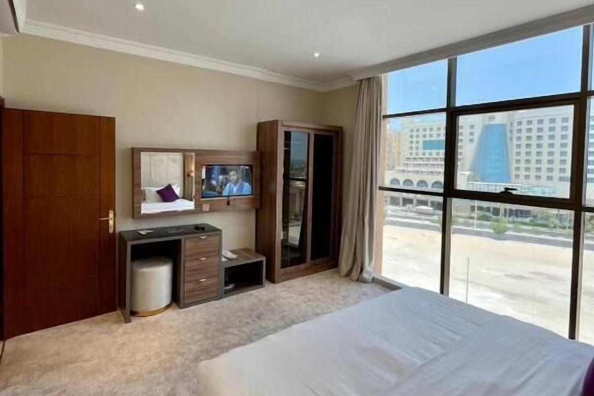 A hotel room with a large bed, a desk with a TV, a wardrobe, and a view of the city.