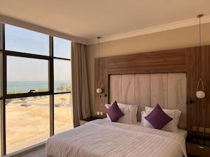 Une chambre d’hôtel dotée d’une grande fenêtre, d’un lit avec une literie blanche et des oreillers violets, d’une tête de lit en bois et d’une table de chevet avec une lampe.