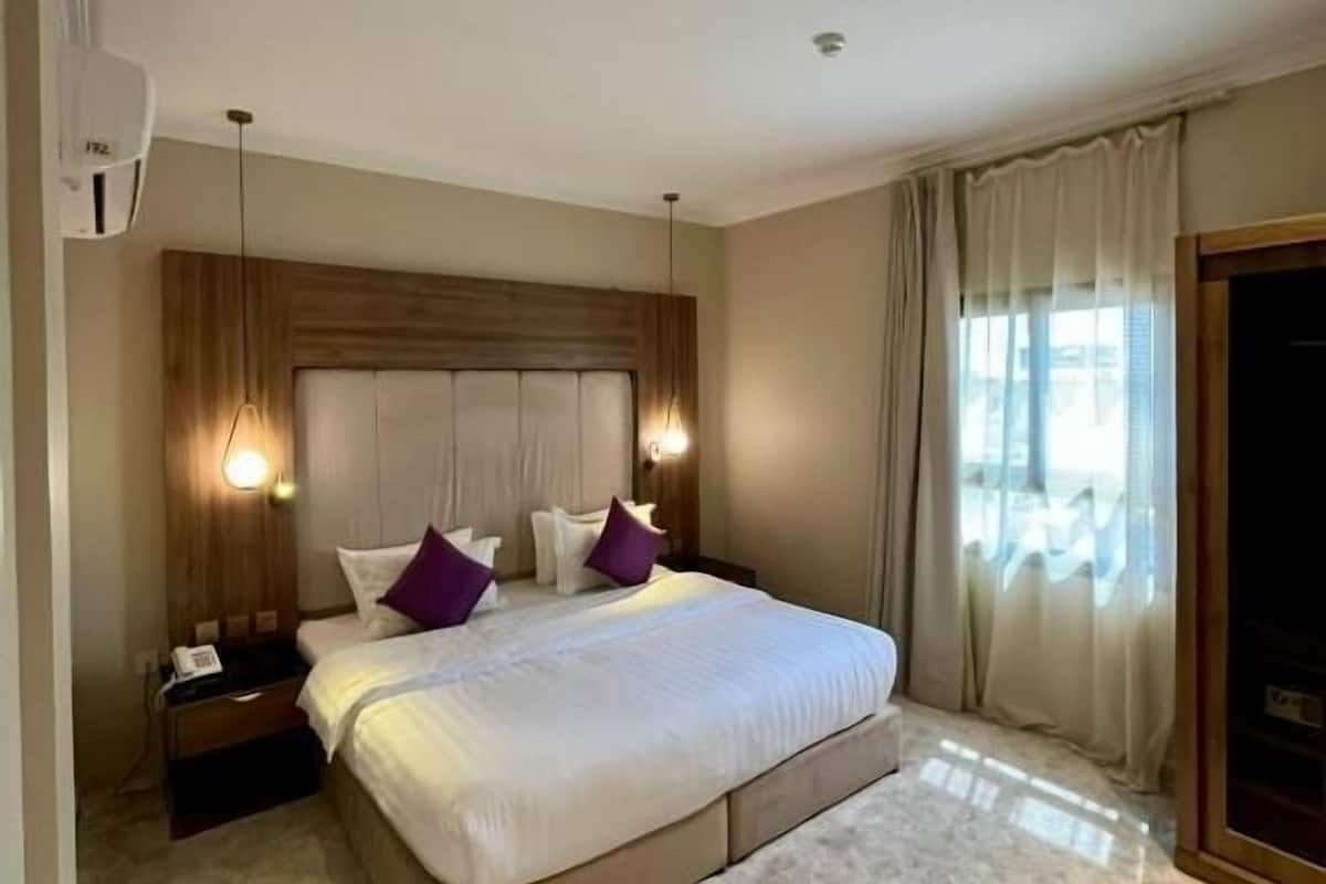Quarto de hotel com cama grande, mesa de cabeceira, luminária na parede, luz no teto e janela com cortinas.