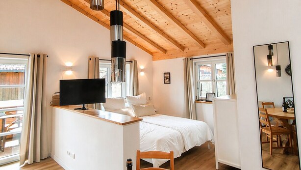 Et moderne interiør med træloft, en central ø med et fladskærms-tv, et spiseområde med bord og stole, samt en seng i baggrunden.