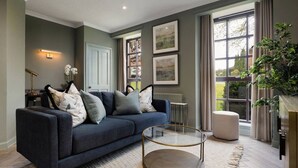 A modern living room with a dark blue sofa, a glass coffee table, and a large window with curtains. - Mill Court Escape & Swimming Pool (Dunblane)