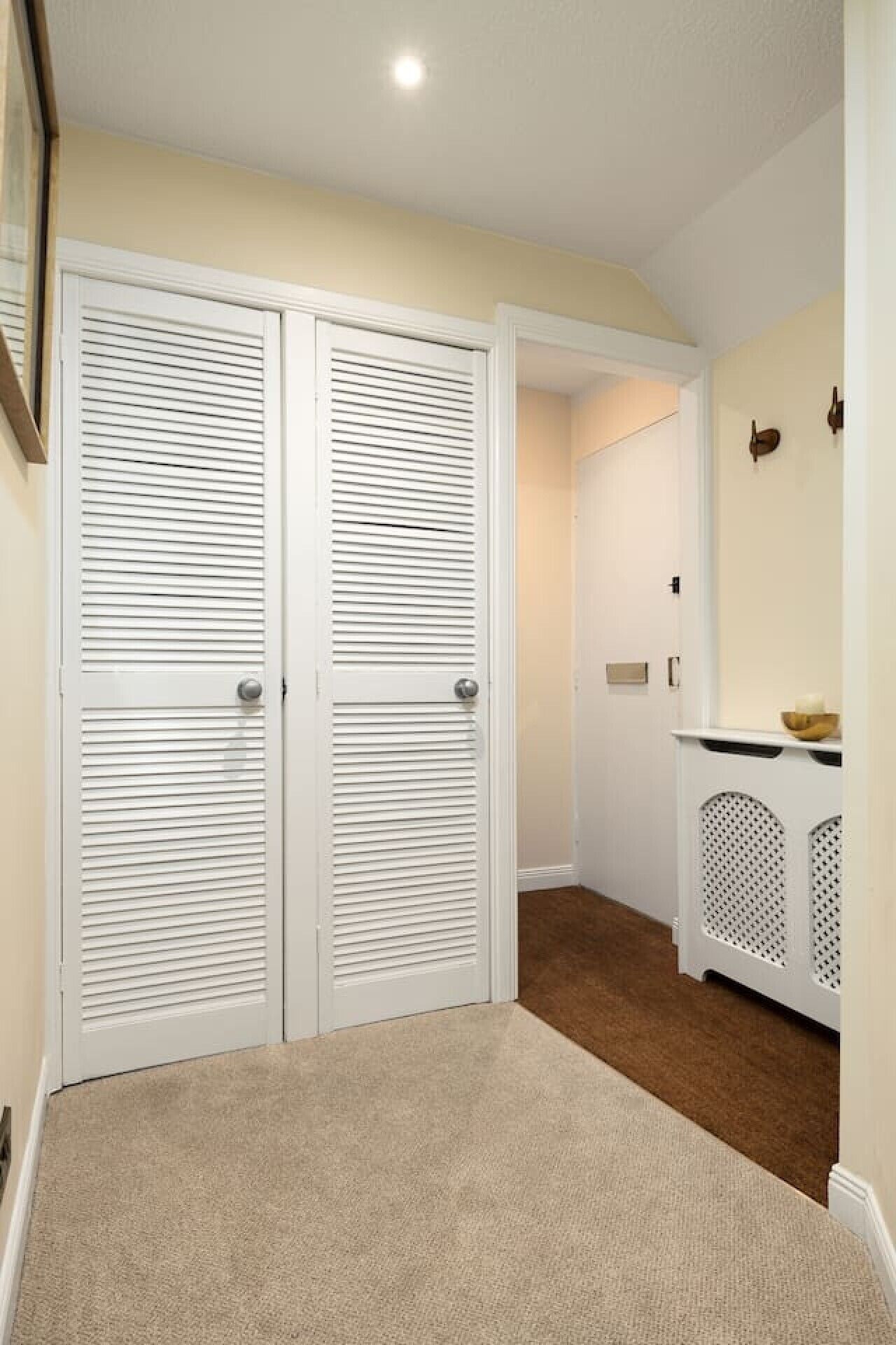 Un pasillo con dos armarios blancos con puertas laminadas, una alfombra beige y una consola blanca con un cuenco y una vela.