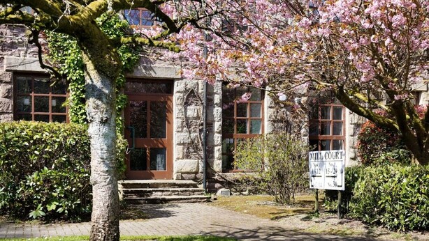 A stone building with a brown door, a signboard, and a tree with pink blossoms. - Mill Court Escape & Swimming Pool (Dunblane)