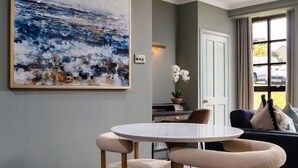 A dining area with a round table, two chairs, a large abstract painting, a potted orchid, and a view through a window. - Mill Court Escape & Swimming Pool (Dunblane)