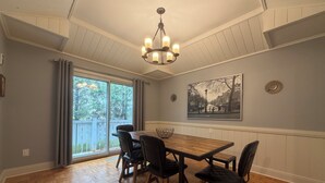 Un coin repas avec une table en bois, quatre chaises, un lustre, une grande fenêtre avec des rideaux et un tableau représentant un paysage urbain accroché au mur.