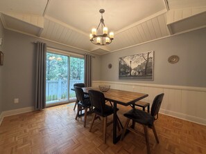 Ein Essbereich mit Holztisch, vier Stühlen, einer Kristallleuchter, einem großen Fenster mit Vorhängen und einem Stadtbild an der Wand.