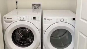 Two white Whirlpool washing machines in a laundry room with a shelf above containing cleaning supplies.