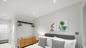 Ein modernes Schlafzimmer mit einem Bett, Nachttisch und einer Wandleuchte. Eine Tür führt in einen anderen Raum, und es gibt ein kleines Holzschränkchen.