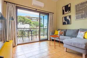 Un salon comprenant un canapé, une petite table, un buffet jaune et une porte-fenêtre coulissante donnant sur un espace extérieur aménagé avec des chaises et une table.