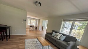 A spacious living area with a sofa, coffee table, and rug, a dining area with chairs, and large windows offering natural light. - Spacious 4 bedroom PRIVATE HOME
centrally located to all the Cape has to offer (Barnstable)