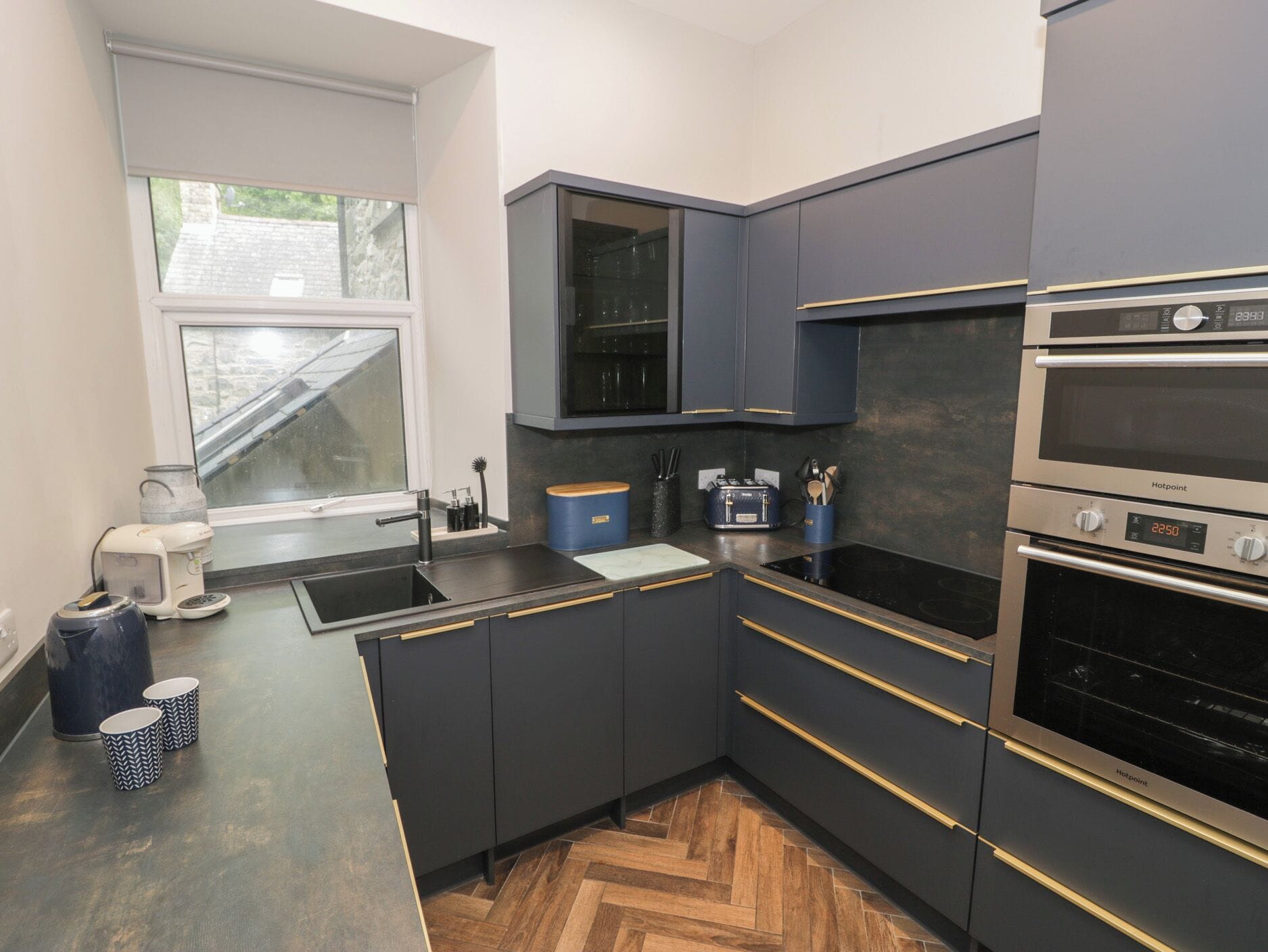 A modern kitchen with dark cabinetry, a built-in oven, and a window overlooking a garden.