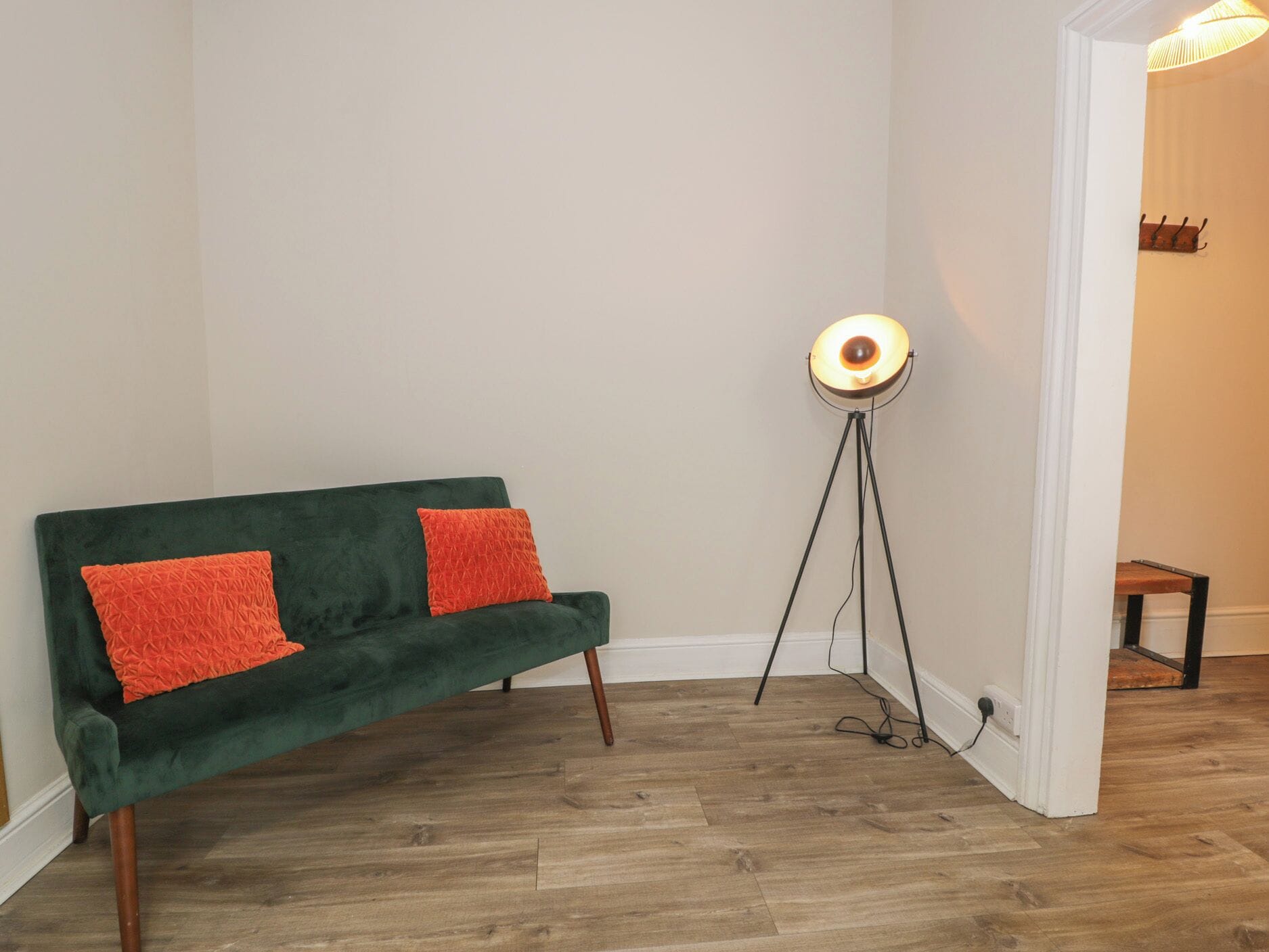 A green sofa with two orange pillows, a floor lamp, and a small wooden stool in a room with wooden flooring and white walls.