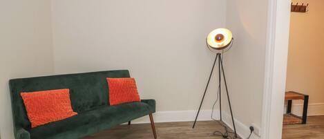 A green sofa with two orange pillows, a floor lamp, and a small wooden stool in a room with wooden flooring and white walls.