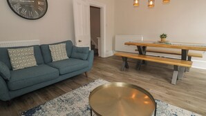 A living room with a blue sofa, a round coffee table, a wooden dining table with benches, a wall clock, and a ceiling light fixture. - Llety Clyd (Barmouth)