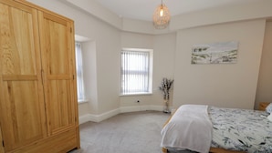 A bedroom with a wooden wardrobe, a bed with a floral bedspread, a window with blinds, and a wall-mounted light fixture. - Llety Clyd (Barmouth)