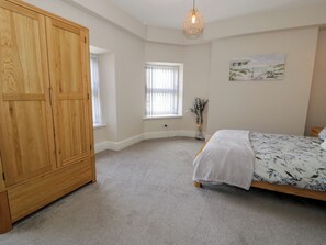 Quarto com guarda-roupa de madeira, cama com colcha florida, janela com persianas e luminária fixada na parede.
