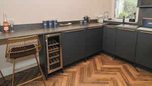 A kitchen with dark cabinets, a built-in wine cooler, a coffee maker, and a window with a view of greenery. - Llety Clyd (Barmouth)