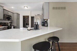 A modern kitchen with a white countertop, stainless steel appliances, and a built-in oven. There are two black stools at the island. - Traditional Meets Design House (Lee`s Summit)