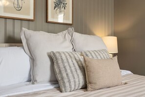 Ein ordentlich bezogenes Bett mit weißer Bettwäsche und drei Kissen, eine Lampe daneben und zwei gerahmte Pflanzenbilder an der Wand.