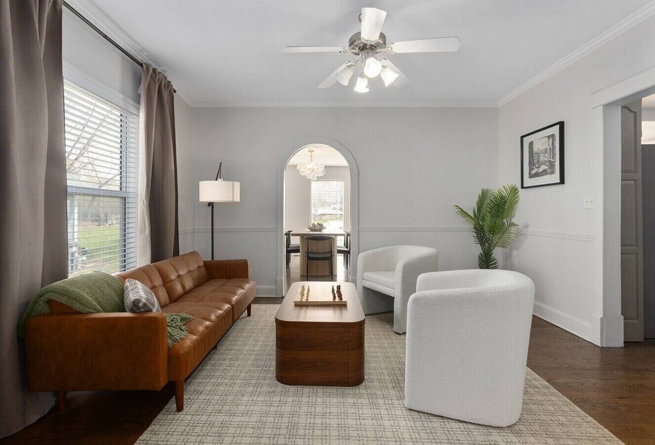 A living room with a brown sofa, a white armchair, a wooden coffee table, a ceiling fan, and a large window with blinds.