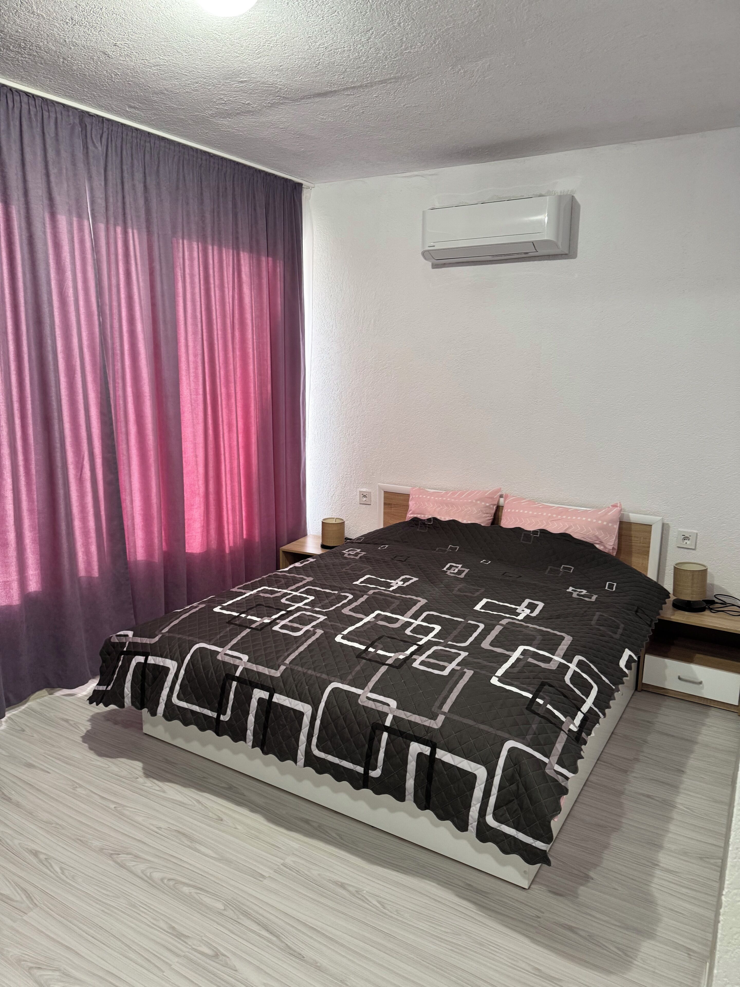 Un dormitorio con cama, cortinas, una mesita de noche y un equipo de aire acondicionado.