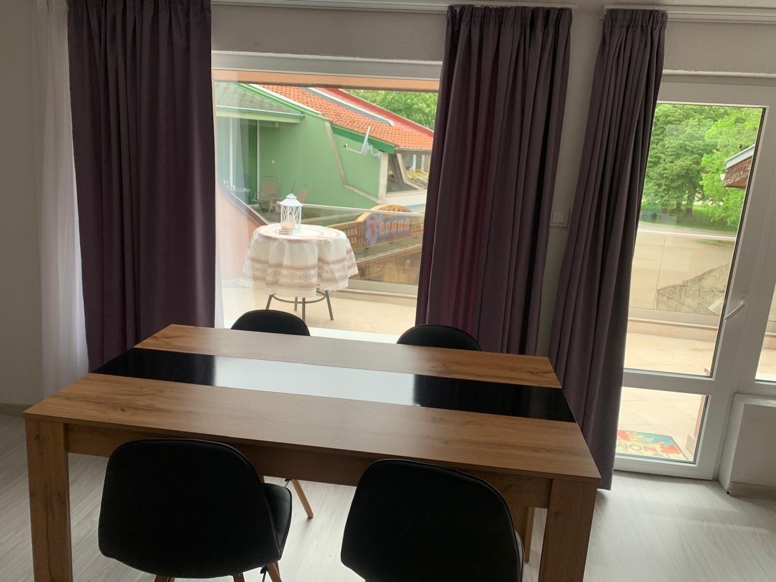 Een kamer met een houten tafel en twee zwarte stoelen, een raam met gordijnen en uitzicht op een groen gebouw.