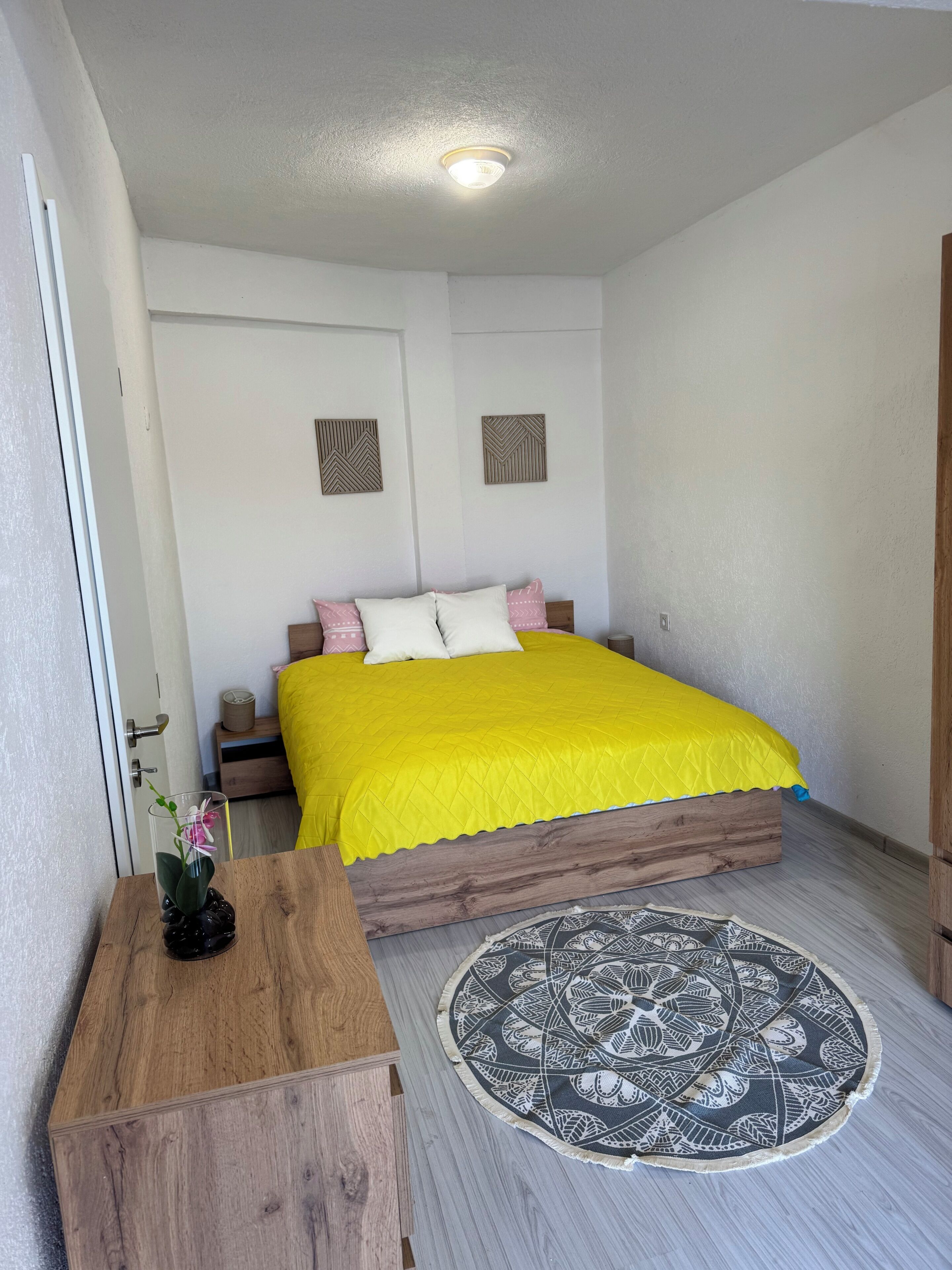 A bedroom with a bed, a wooden nightstand, a bedside table, a vase with flowers, a patterned rug, and a door.