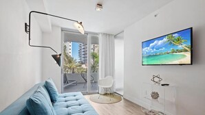 A modern living room with a sofa, a TV mounted on the wall, a clear glass cabinet, and a sliding glass door leading to a balcony with outdoor furniture. - 6080 Collins  (Miami Beach)