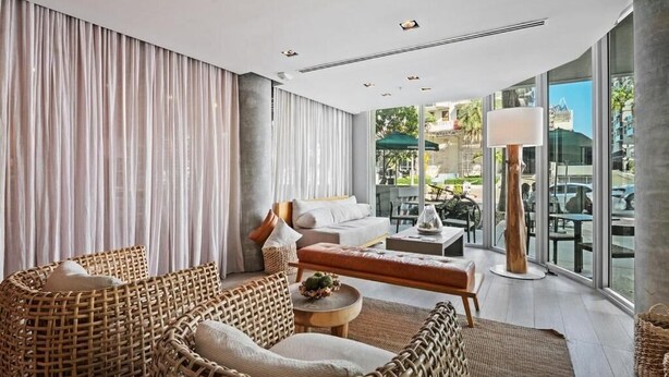 A modern hotel room with a large window, wicker furniture, a sofa, a round coffee table, and a floor lamp. - 6080 Collins  (Miami Beach)