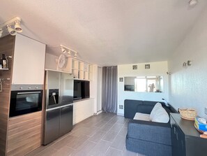 A compact kitchen with a built-in oven, refrigerator, and microwave. There is a grey sofa and a small table with a basket. - Cosy studio with sea & garden view - swimming pool (Sète)