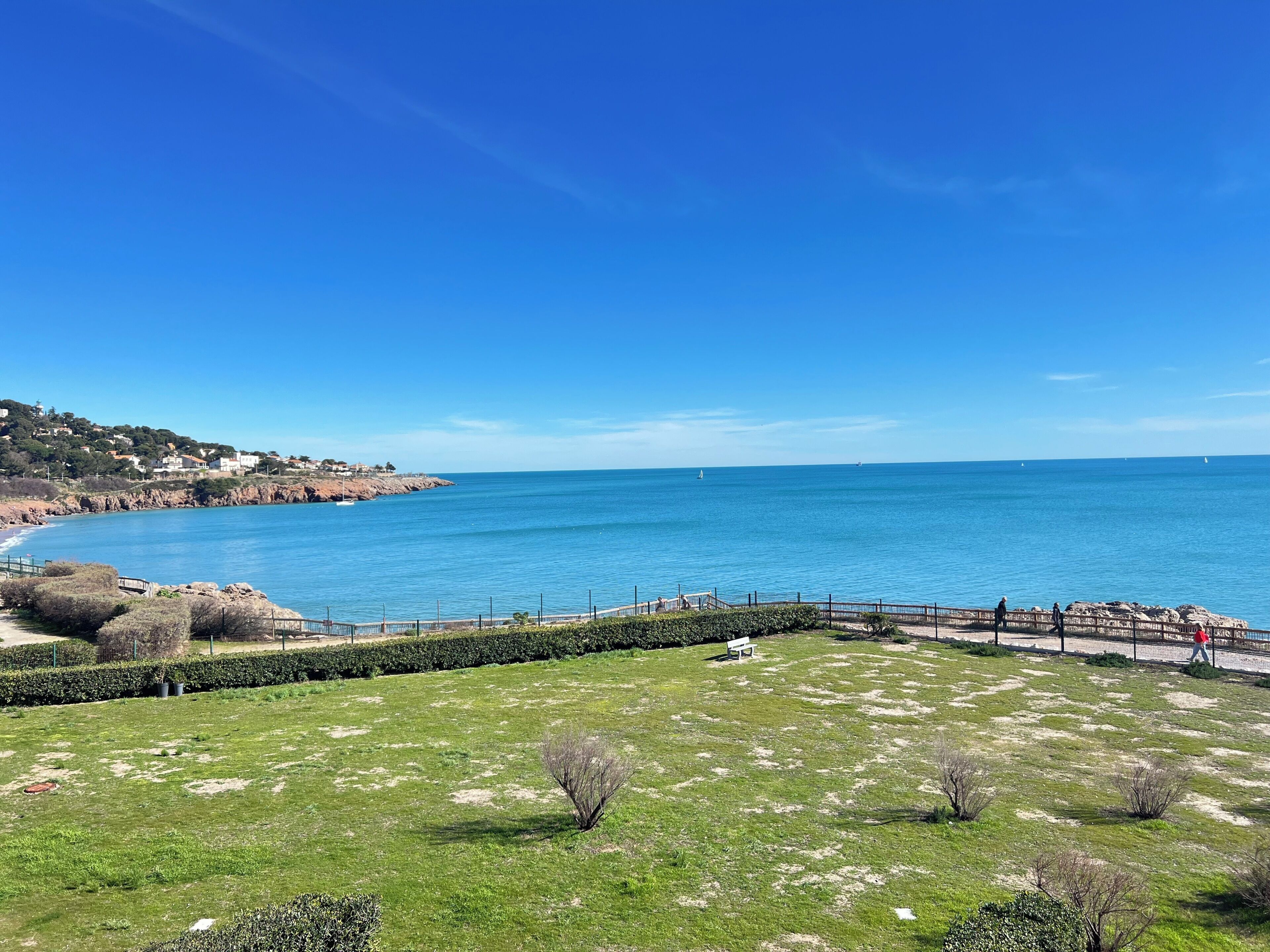 A coastal landscape with a clear sky, a grassy area with a bench, and a view of the sea.